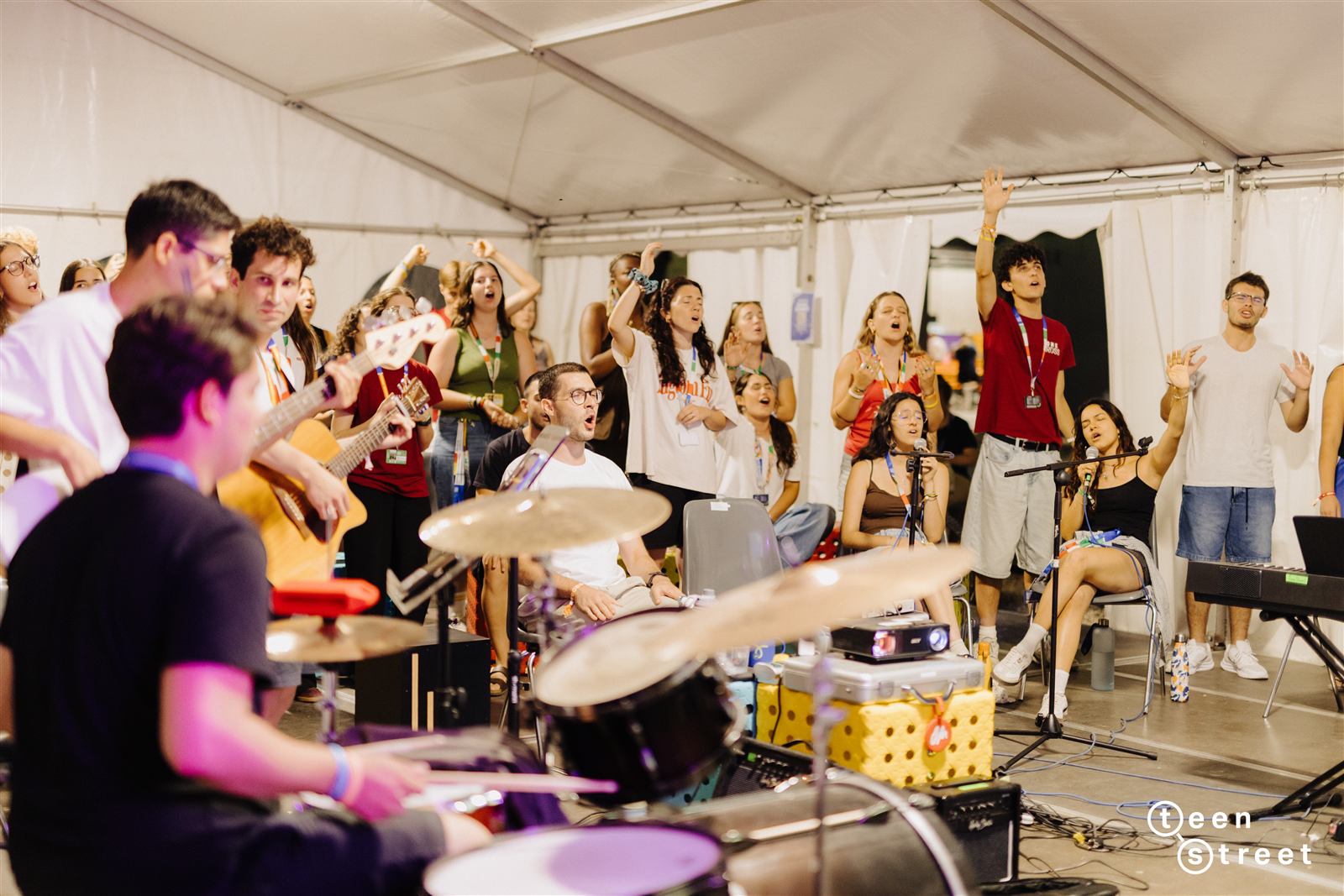 Pequeno grupo a louvar numa tenda, com guitarra, bateria e adolescentes de braço no ar